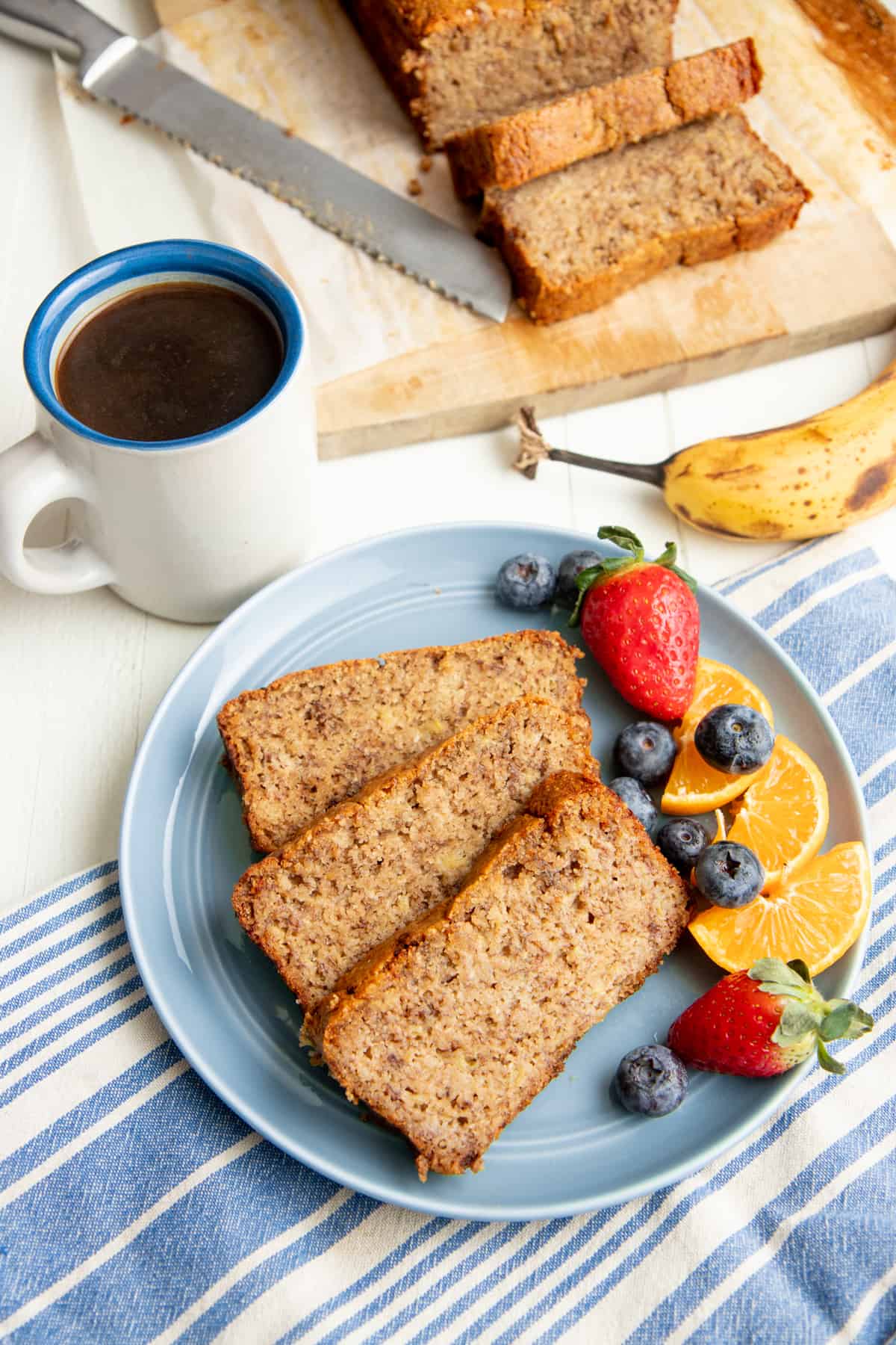 Three slices of paleo banana bread on a blue plate with mixed fruit, next to a cup of coffee.