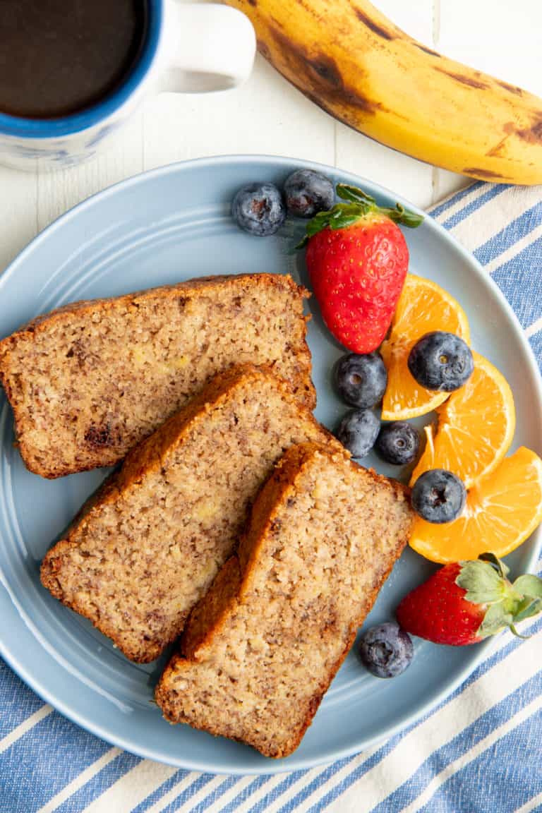 Three slices of almond flour banana bread on a blue plate with mixed fruit, next to a cup of coffee.