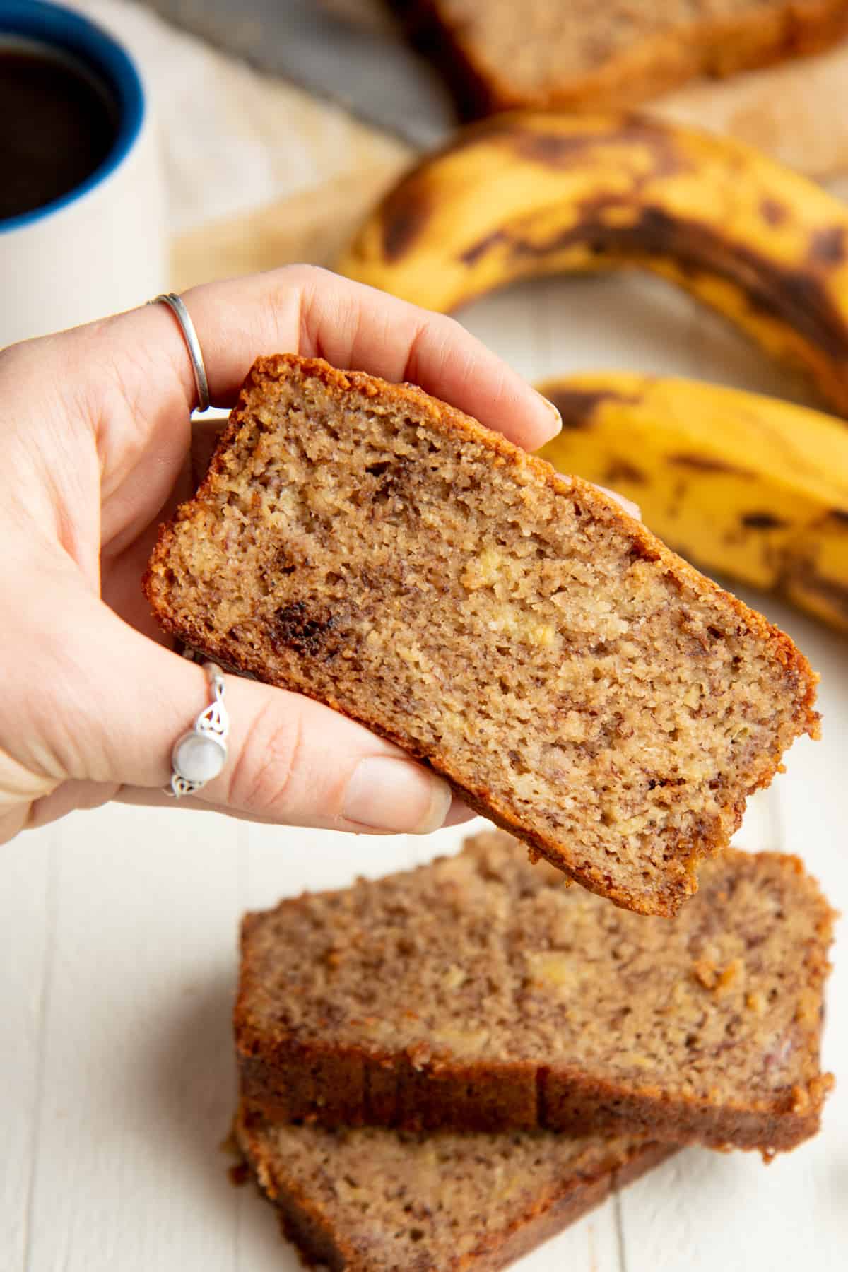 A woman's hand holds a slice of almond flour banana bread over a stack of other slices.