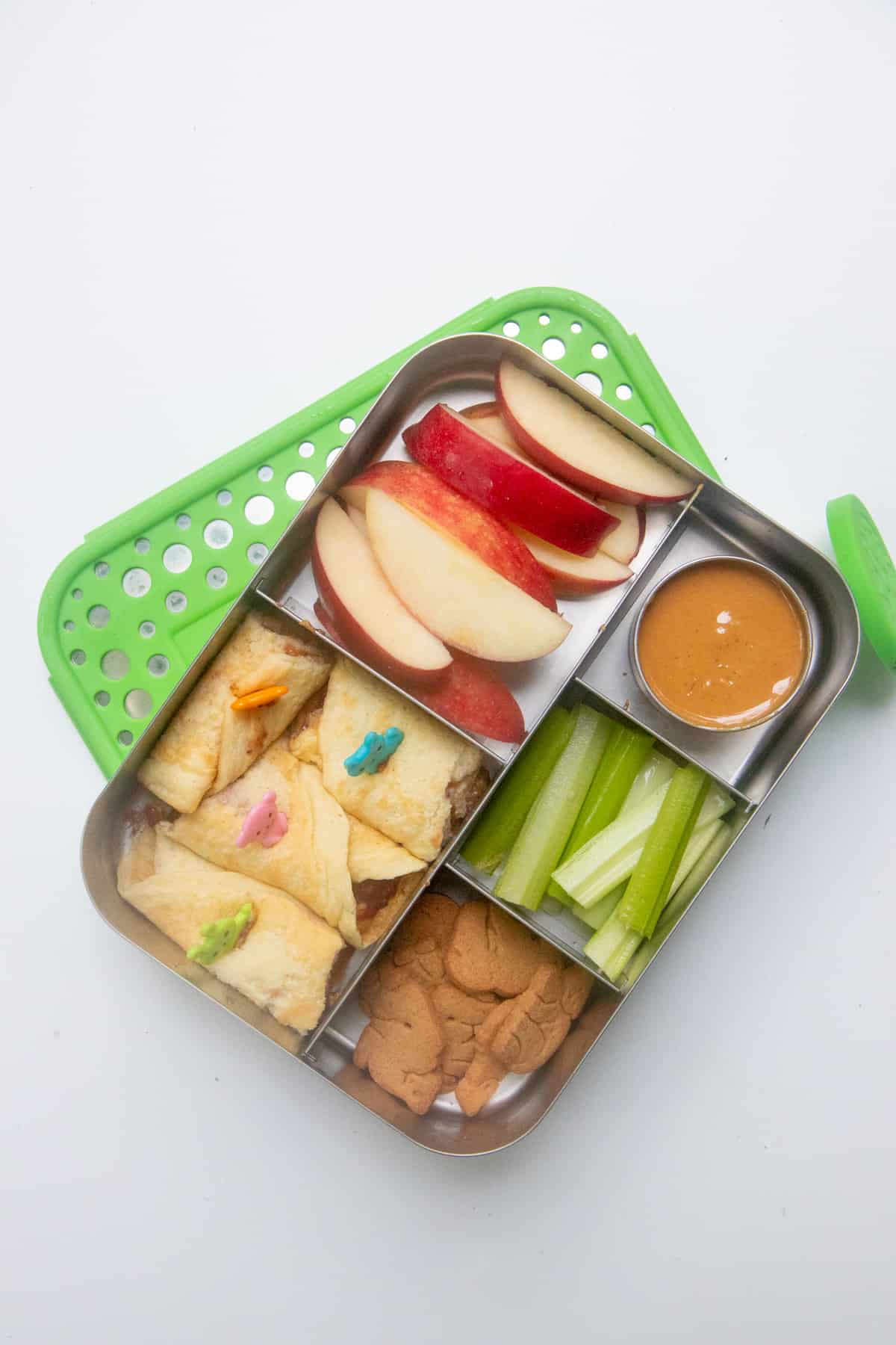 Stainless steel lunch box with pancake roll-ups, apple slices, celery, and animal crackers.