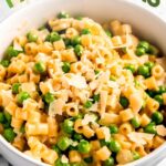 Creamy ditalini pasta with peas, garnished with shaved parmesan in a white bowl on a light counter. A text overlay reads, "Pasta & Peas."