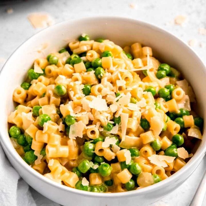 Creamy ditalini pasta with peas, garnished with shaved parmesan in a white bowl on a light counter.