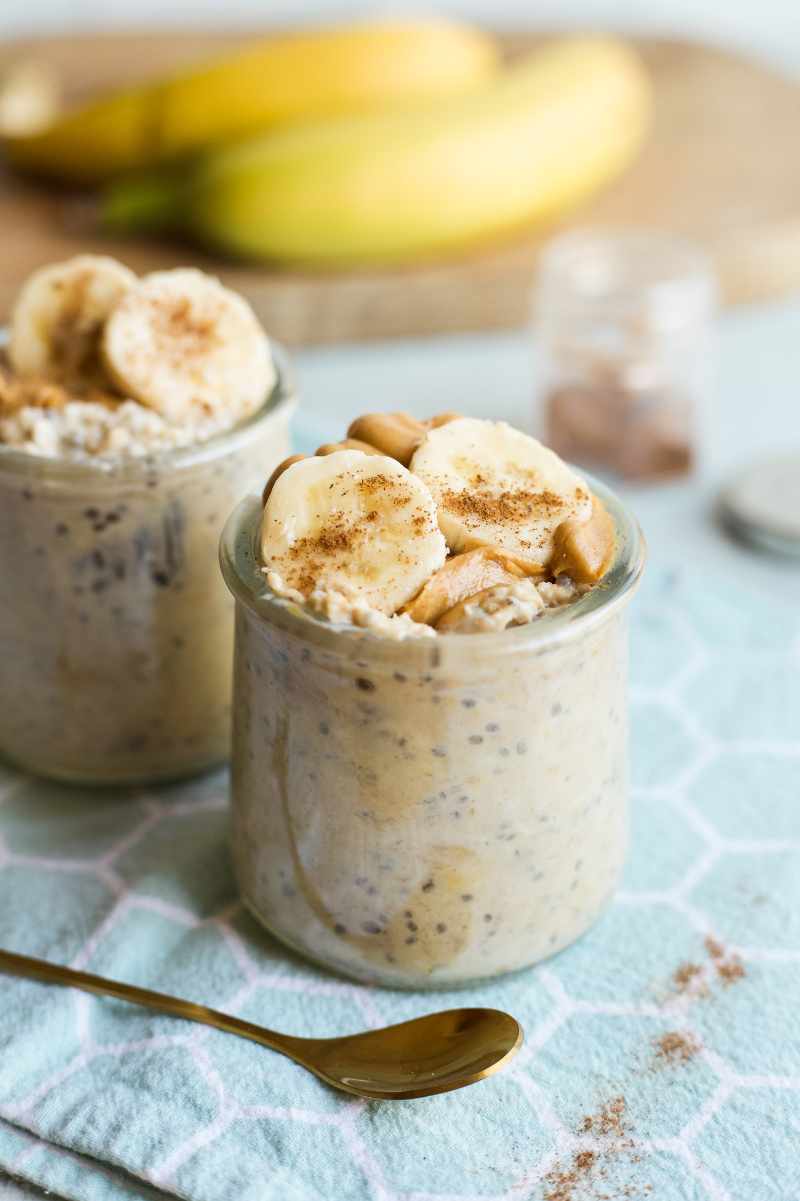 Two jars of overnight oats topped with peanut butter and banana slices sprinkled with cinnamon stand on a honeycomb pattern kitchen linen.