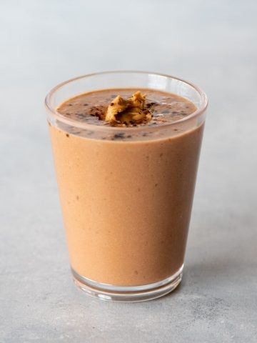 A peanut butter smoothie topped with a scoop of peanut butter and a sprinkling of cocoa powder in a glass on a light background.