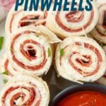 Close view of pepperoni pinwheel bites arranged on a plate around a small bowl of marinara sauce. A text overlay reads, "Pepperoni Pinwheels."