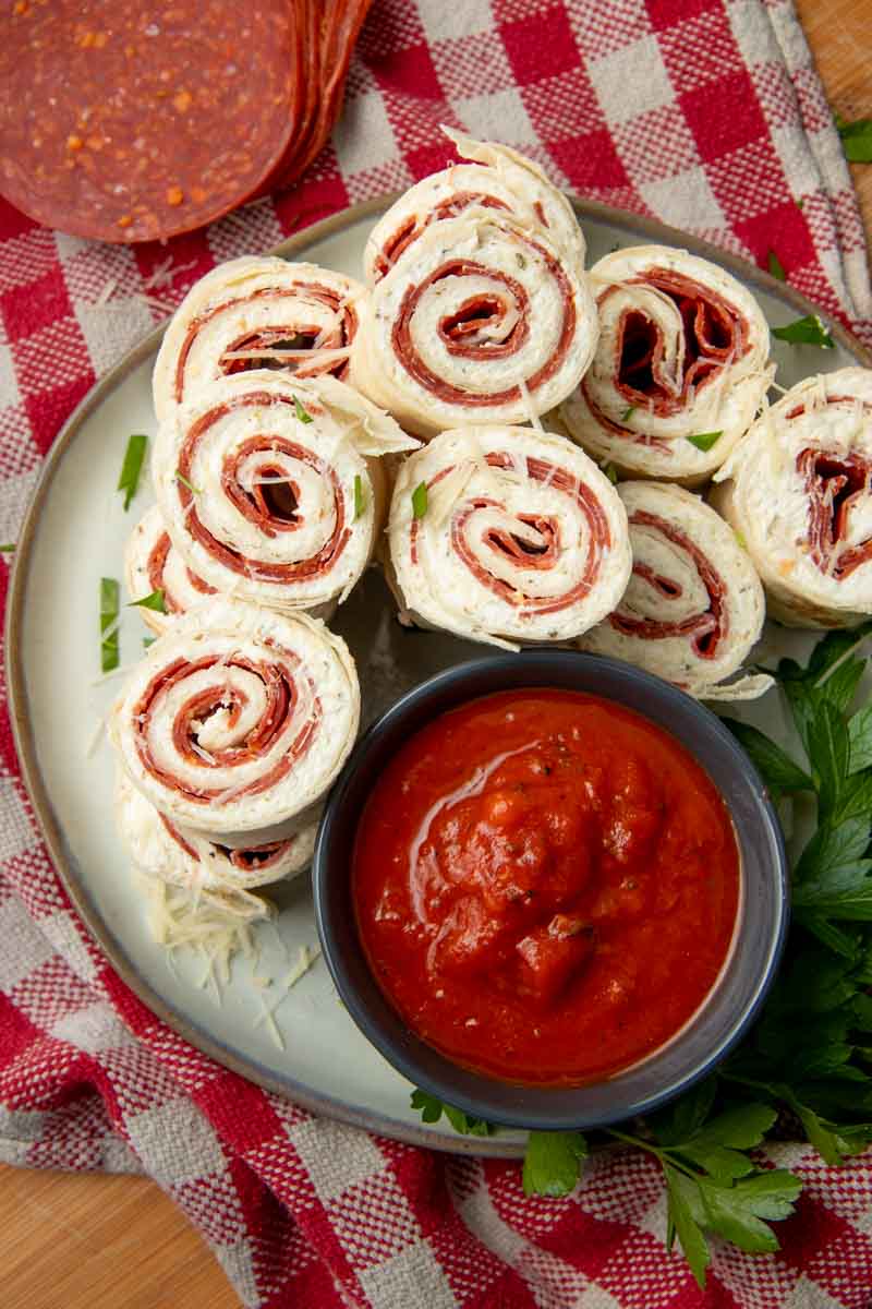 Overhead of a party platter of pizza roll-ups garnished with shredded parmesan cheese and italian flatleaf parsley, a bowl of marinara sauce nestled alongside for dipping.