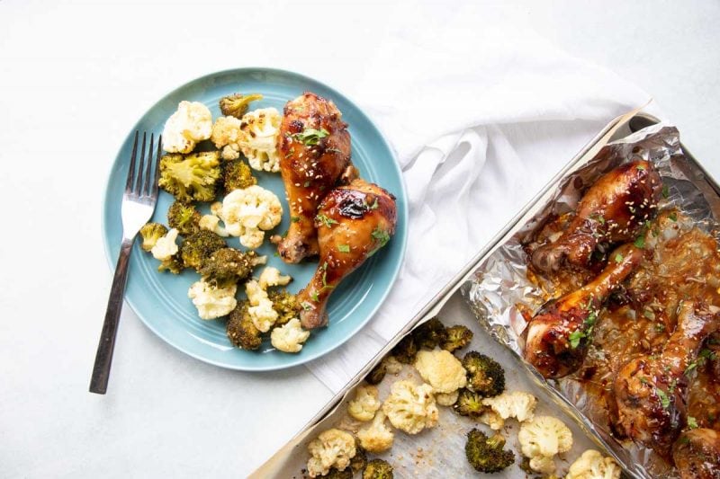 A dinner platter of baked chicken legs and vegetables sits beside a foil lined sheet pan with the remaining drumsticks and vegetables.