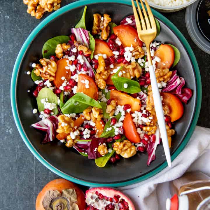 A fork rests on a dark bowl, filled with persimmon and pomegranate salad.