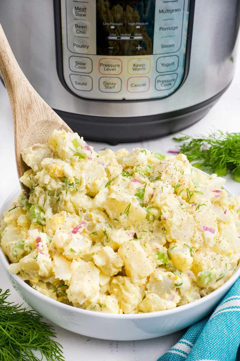 A wooden spoon scoops into a bowl of potato salad sitting in front of an instant pot multi-cooker.