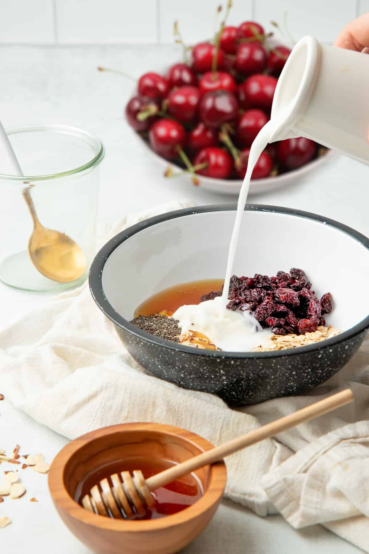 A pitcher pours milk into a mixing bowl full of oats, Greek yogurt, chia seeds, dried cherries, and other ingredients.