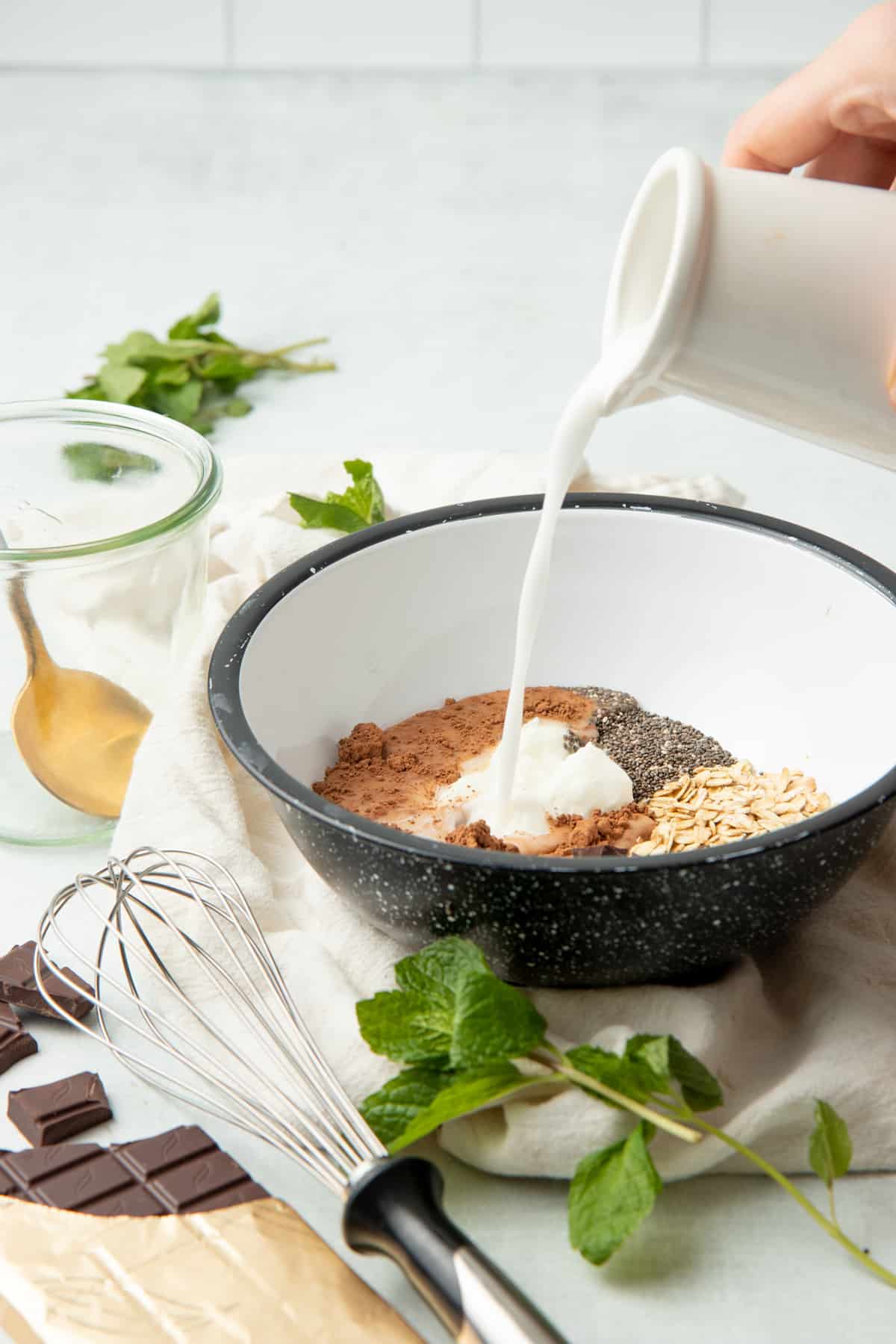 A hand pours milk into a bowl of overnight oat ingredients. A partially unwrapped sits in the bottom left corner.