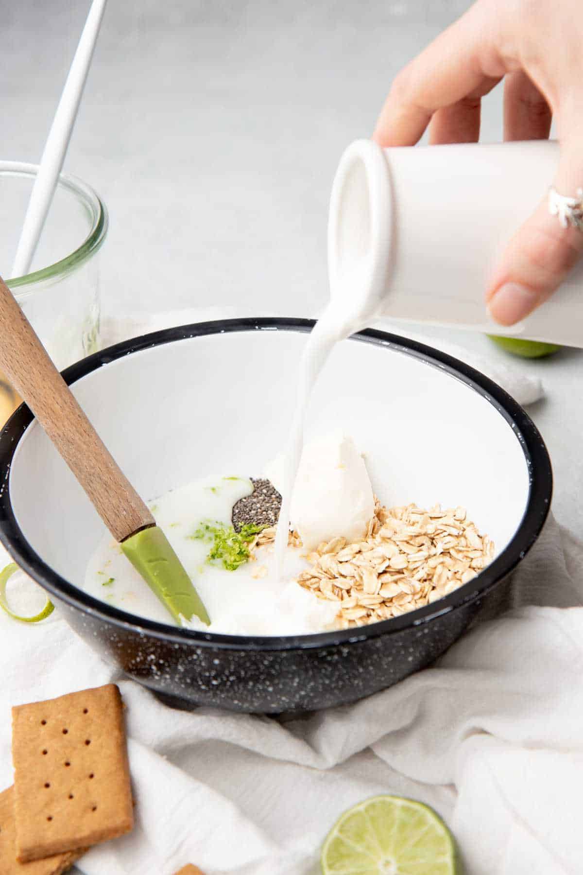 A hand pours milk into a white bowl of overnight oat ingredients.