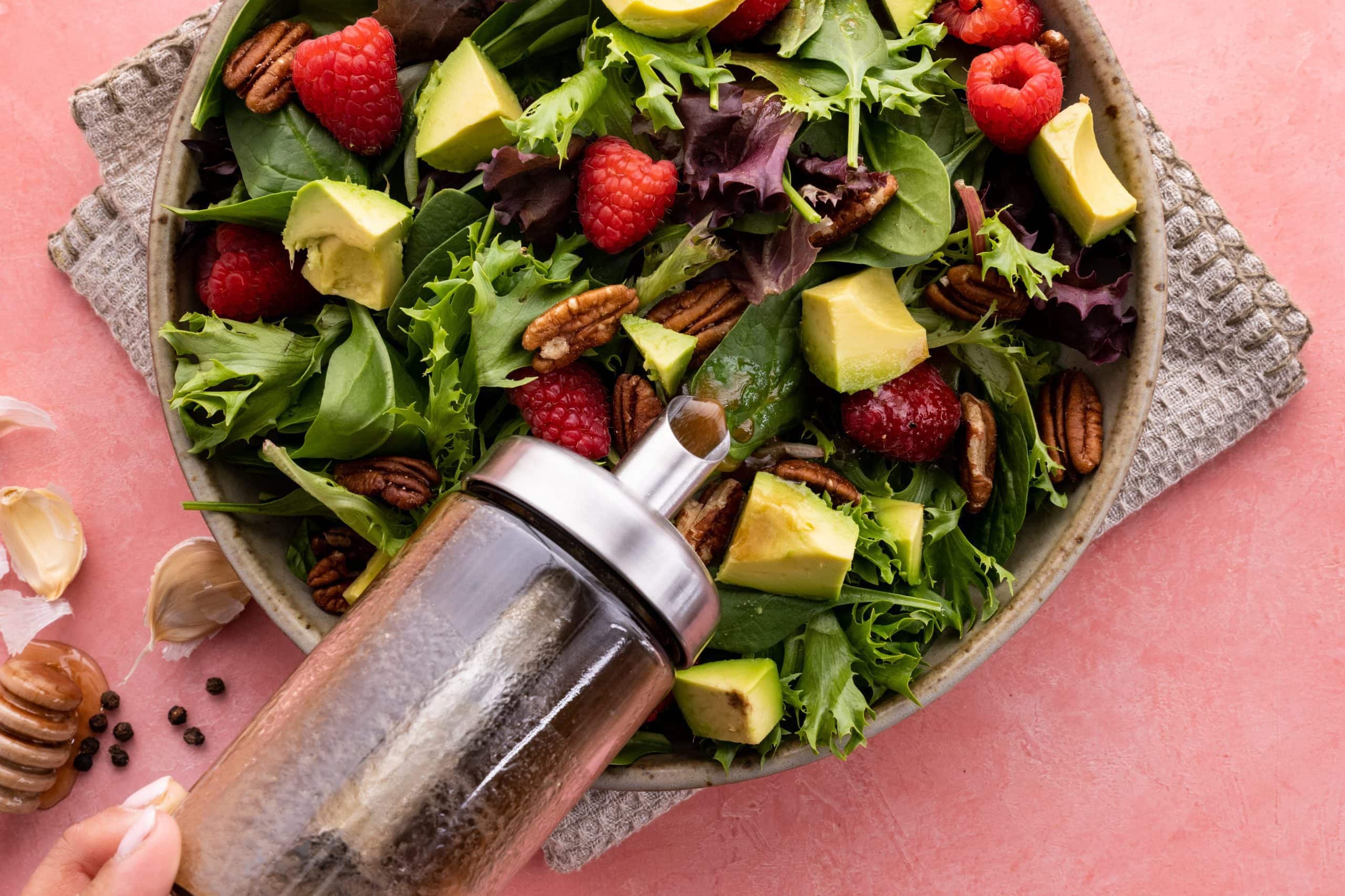 Top view of a hand pouring vinaigrette on a salad of fresh greens, raspberries, pecans, and avocado.