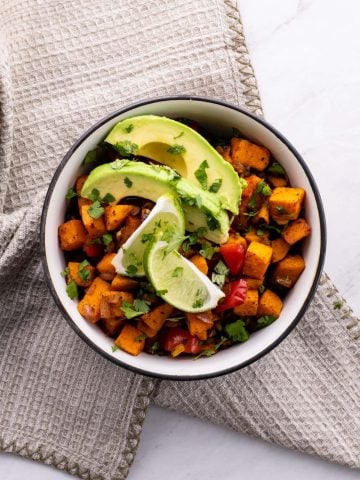 Two black-rimmed bowls filled with sweet potatoes, lime slices, avocado slices, black beans, and rice.