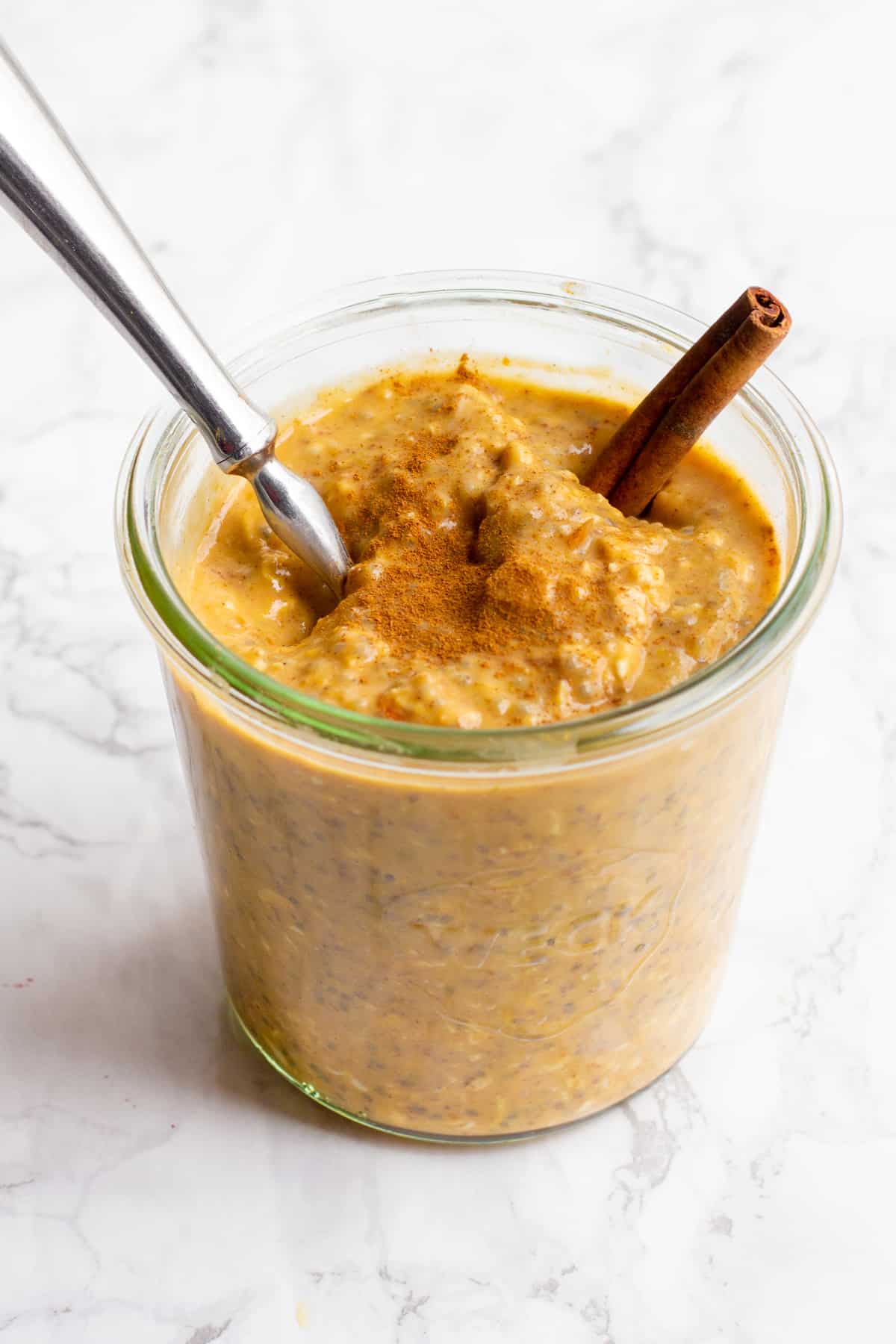 A glass jar filled with pumpkin spice overnight oats sits on a marble countertop. The oats are garnished with a cinnamon stick, and a spoon dips into the jar.