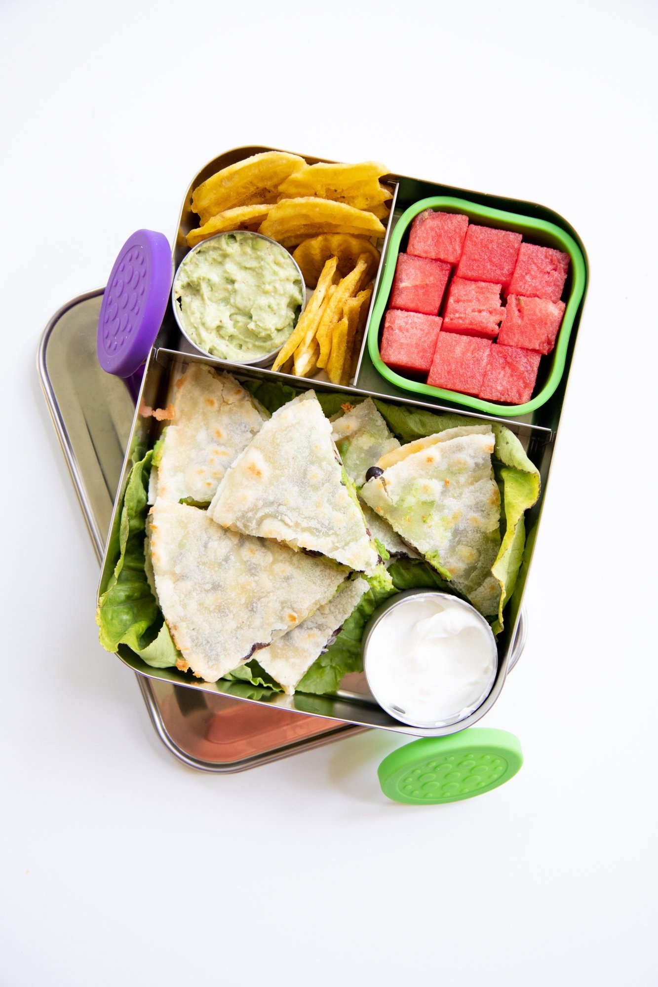 Close up on a metal bento box with avocado black bean quesadilla, chips, guacamole, and watermelon