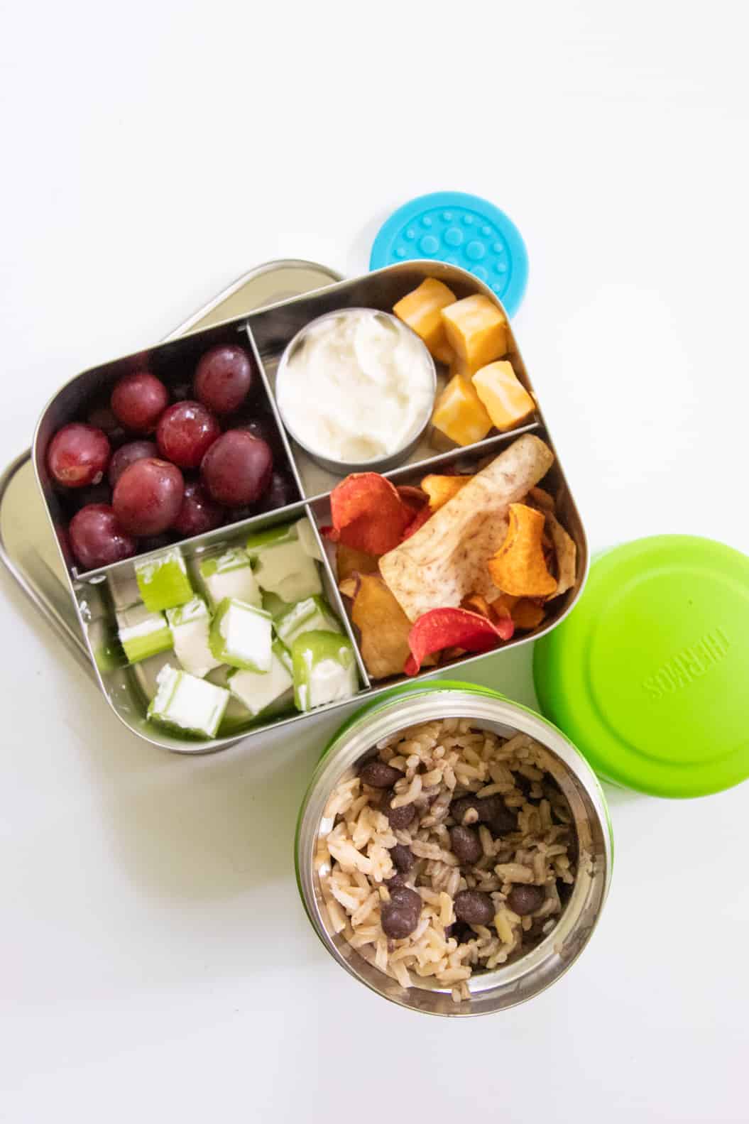 Stainless steel lunch box with grapes, cheese, ants on a log, and veggie chips, next to a Thermos of rice and beans