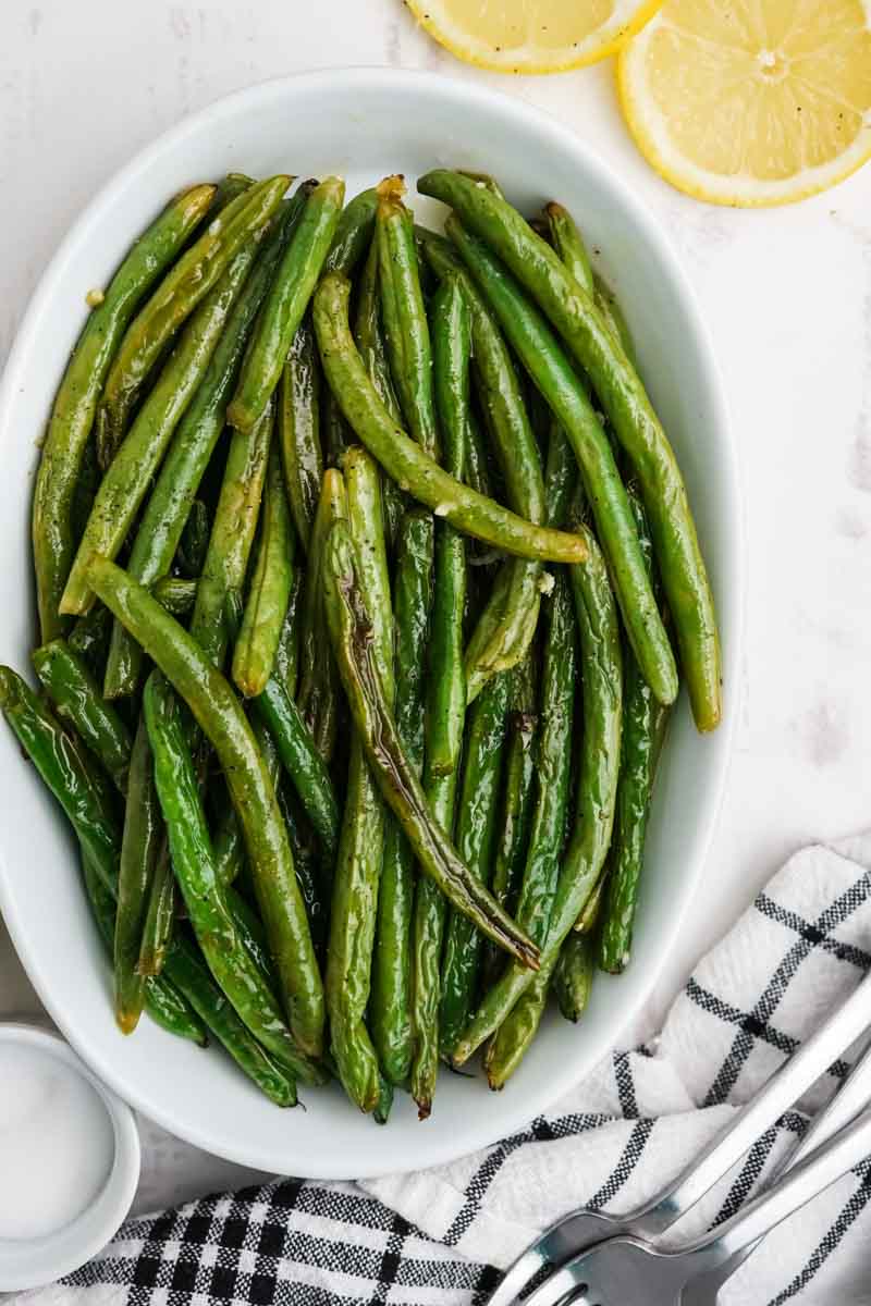 Overhead view of roasted green beans in a white serving dish with a cloth napkin and forks nearby.