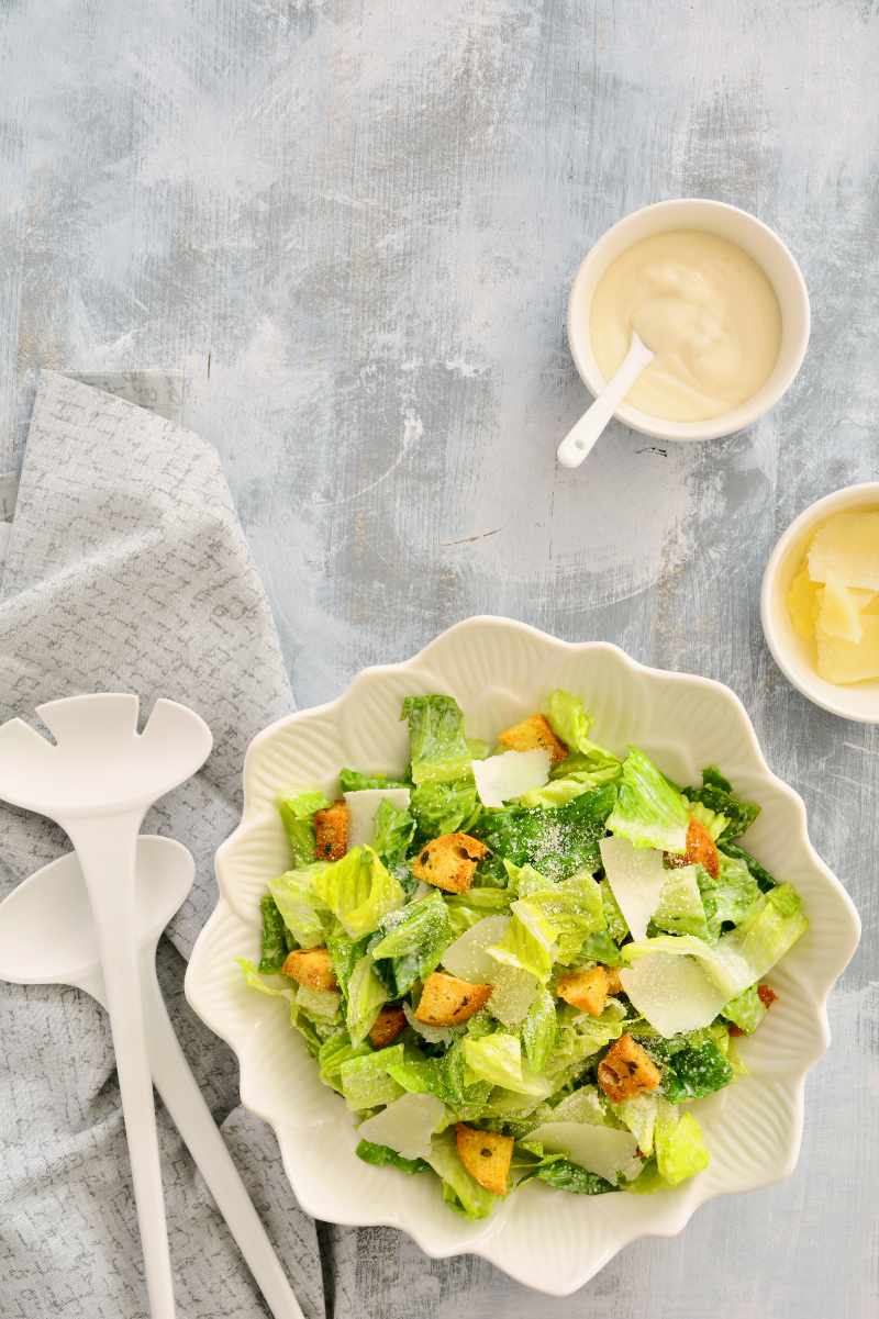 Traditional caesar salad in a white bowl with salad servers and two small bowls of dressing and shaved parmesan alongside.