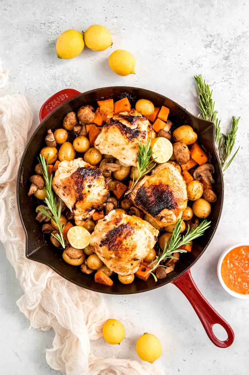 Overhead of a red enameled Staub cast iron skillet with roasted baby potatoes, button mushroom halves, and sweet potato cubes in the bottom and four lemon rosemary chicken thighs nestled on top.