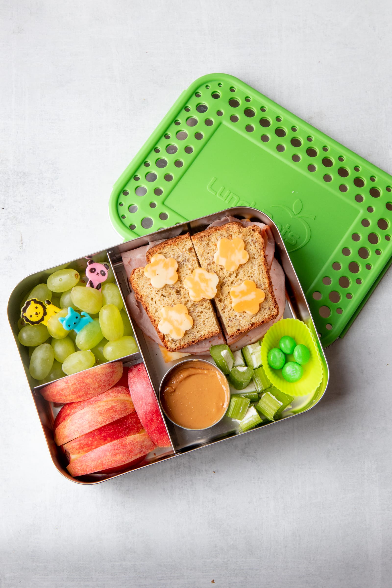 A zero-waste packed lunch consisting of a divided lunch box with a sandwich, grapes, apple slices, and celery.