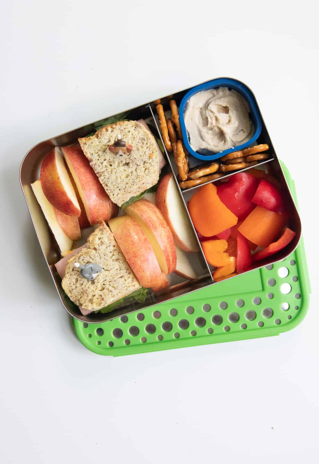 Stainless steel lunch box with a sandwich, apple slices, peppers, and pretzels with hummus.