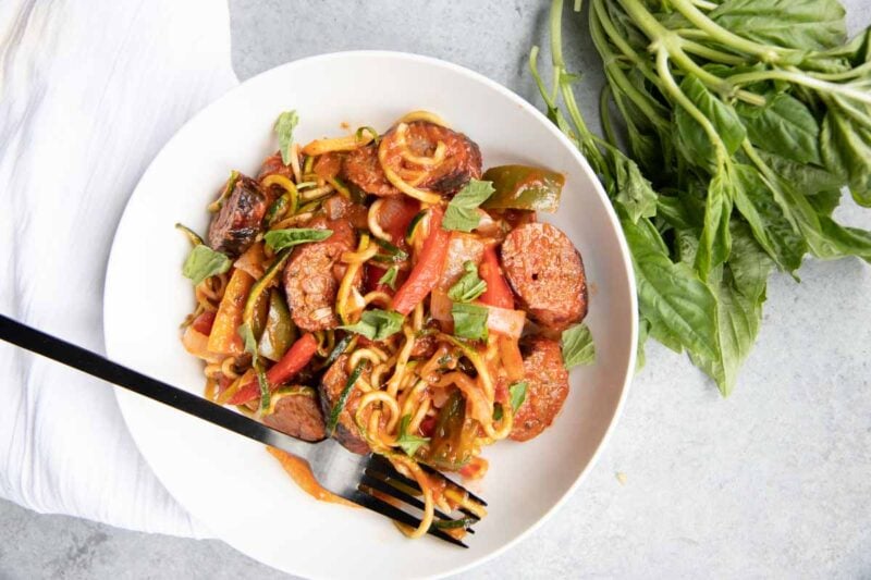 A sausage and veggies dinner plated on a white dish with a dark metal fork. A bunch of fresh basil lies next to the plate.