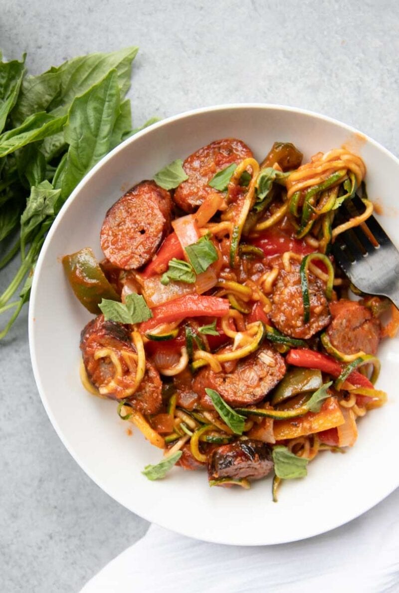 A saucy sausage zoodle dish on a white plate with fresh basil garnish.