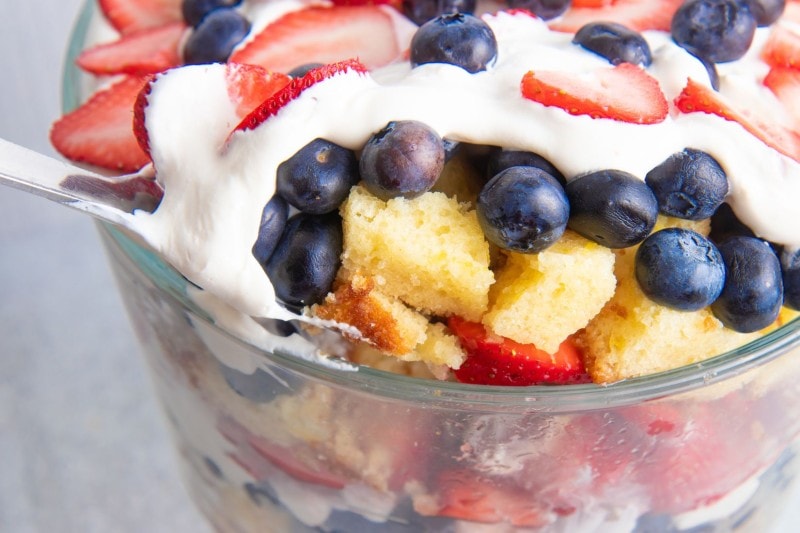 A large serving spoon scoops berry trifle from a large straight-sided glass serving dish.