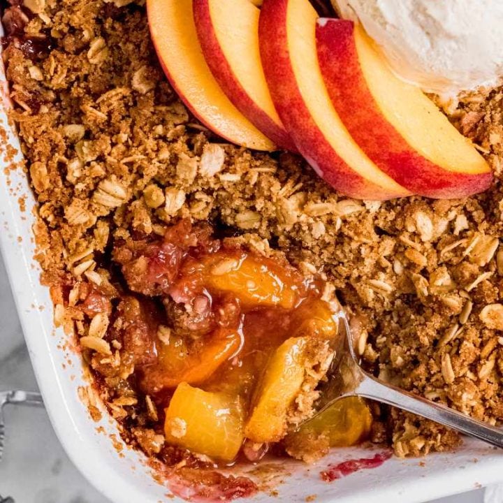 Overhead of a spoon dipping into a peach crisp. Fresh peach slices are arranged on the top of the middle of the crisp.