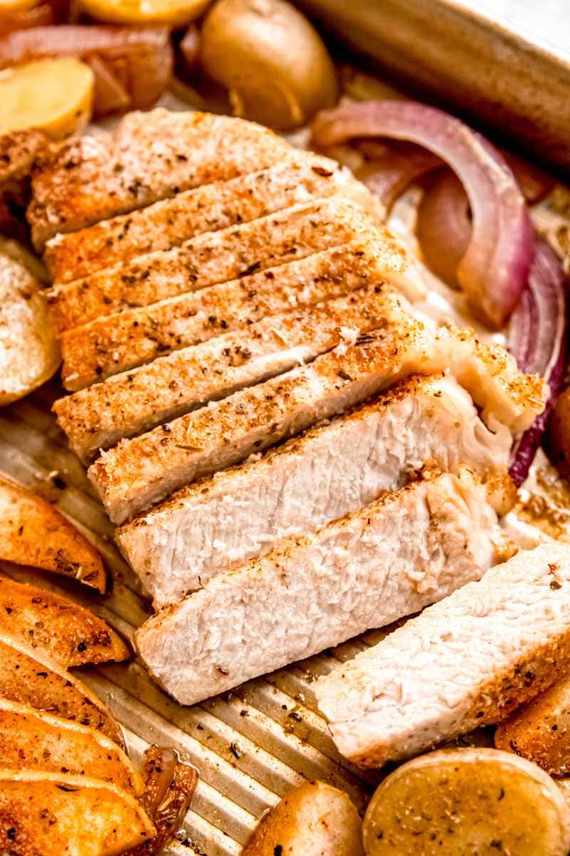 A seasoned pork chop sliced on a pan with onions, apples, and potatoes.