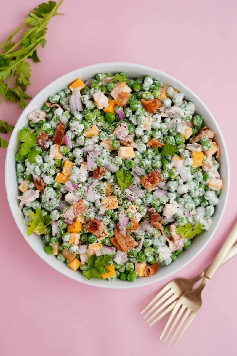 A large white serving bowl of creamy pea salad with cheese and bacon rests on a pale pink counter with forks nearby.