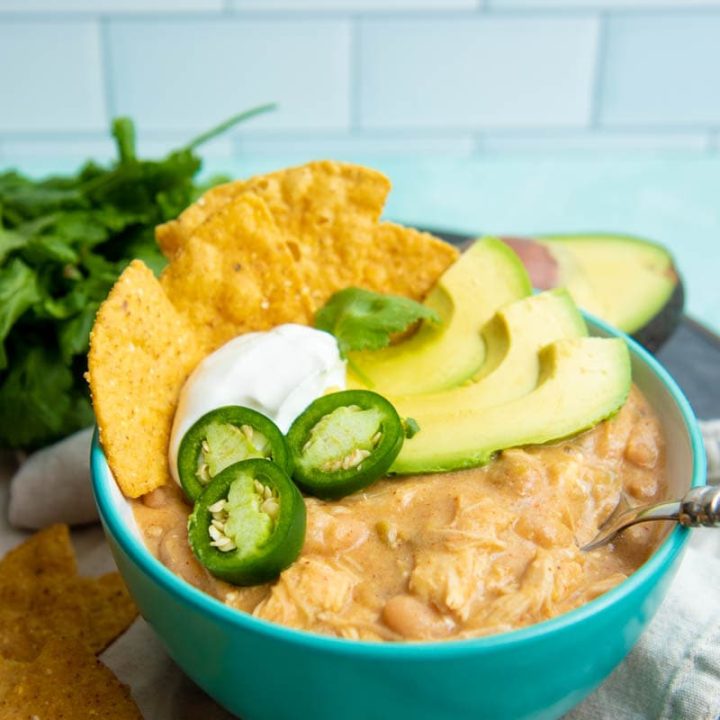 A teal bowl filled with Instant Pot white chili topped with sour cream, avocado, jalapeno, and corn tortilla chips.