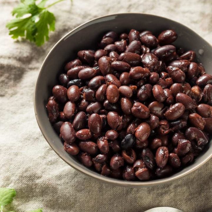 A bowl of instant pot black beans on a linen table cloth with a spoon and sprigs of fresh herbs nearby.