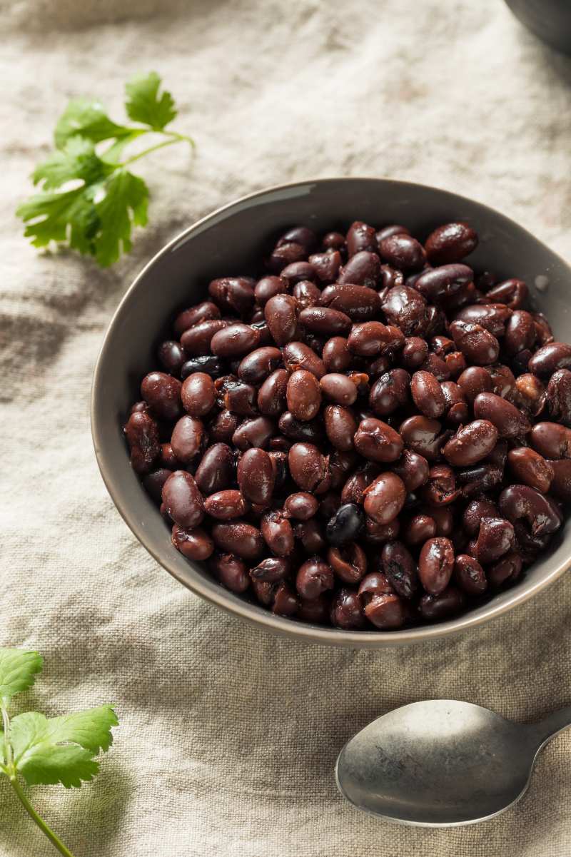 A bowl of instant pot black beans on a linen table cloth with a spoon and sprigs of fresh herbs nearby.