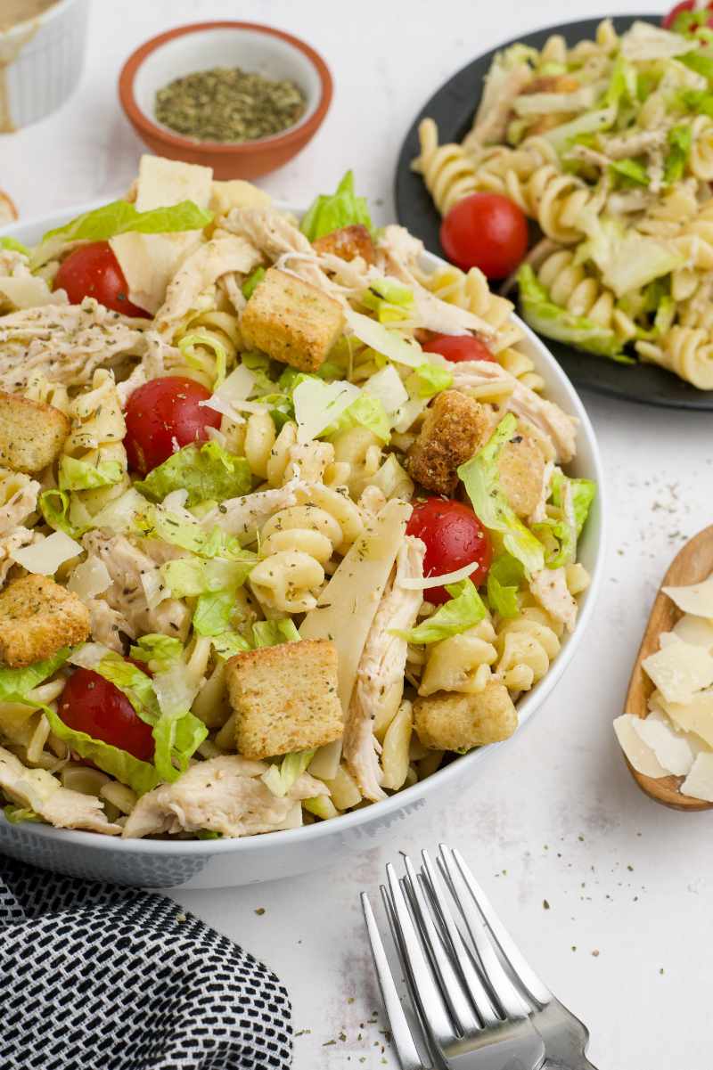 A large white serving bowl of pasta sits on a light countertop with forks and garnishes alongside.