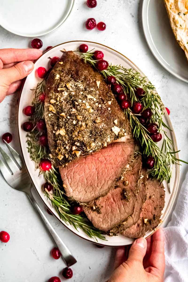 Two hands set a platter of roast beef on a light table with a large serving fork alongside.