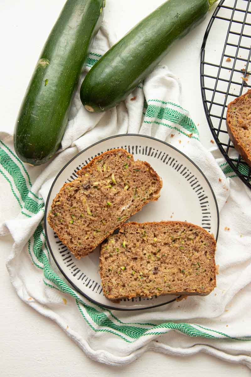 Two slices of gluten-free zucchini bread served on a white plate atop a kitchen linen with a green stripe, two whole zucchini nearby.