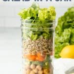 A tall mason jar with layered salad ingredients sits on a white counter in front of a white wall. Lemon slices sit next to the jar in a small black bowl. A text overlay reads "Sesame Chickpea Salad in a Jar."