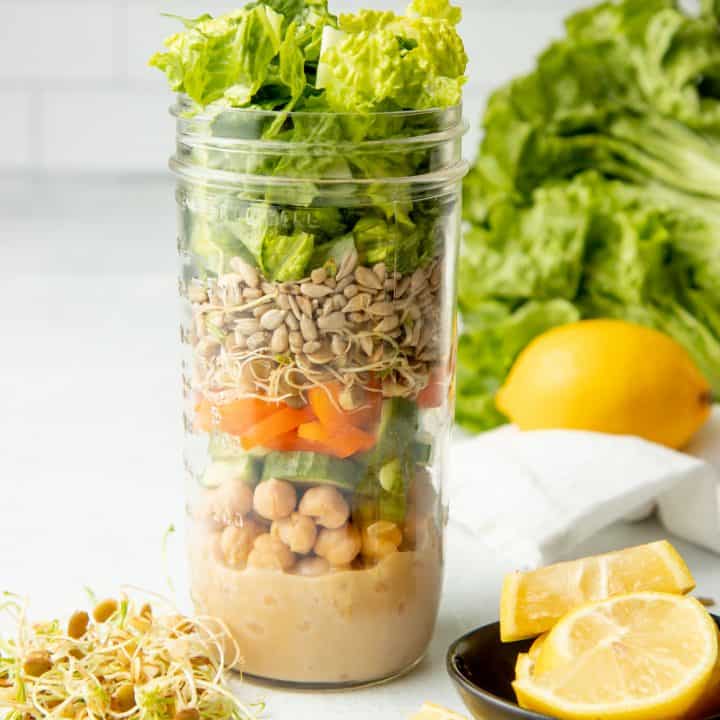 A tall mason jar with layered salad ingredients sits on a white counter in front of a white wall. Lemon slices sit next to the jar in a small black bowl.