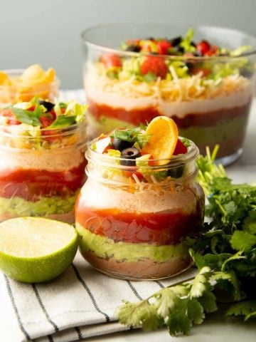 Two individual servings of seven layer dip stand on a kitchen linen in front of a party size bowl of dip.