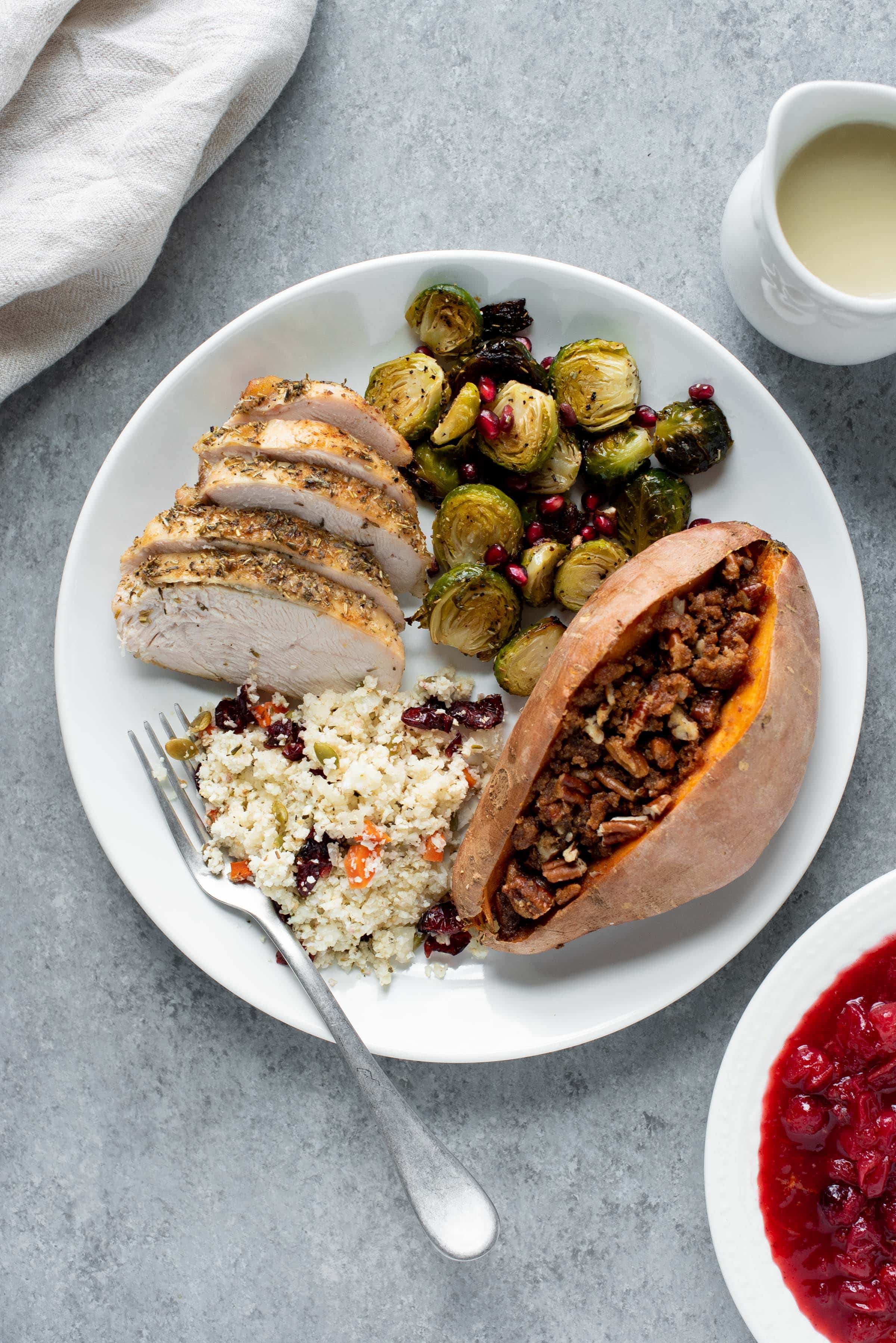 Paleo Thanksgiving dinner on a white plate with a fork. Components include sweet potatoes, Brussels sprouts, turkey, and cauliflower rice pilaf