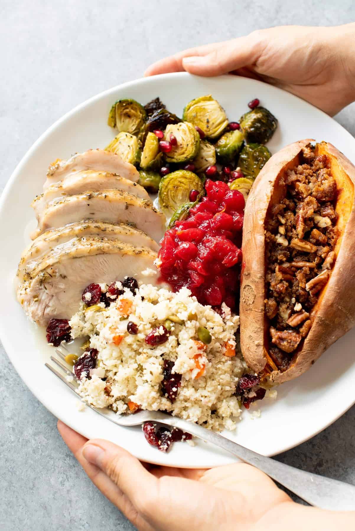 Hands holding a plate of Thanksgiving food - turkey, Brussels sprouts, cranberry sauce, sweet potatoes, and cauliflower rice pilaf