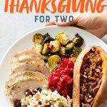 Hands holding a plate of Thanksgiving food - turkey, Brussels sprouts, cranberry sauce, sweet potatoes, and cauliflower rice pilaf. Text overlay reads "Sheet Pan Paleo Thanksgiving Dinner for Two."