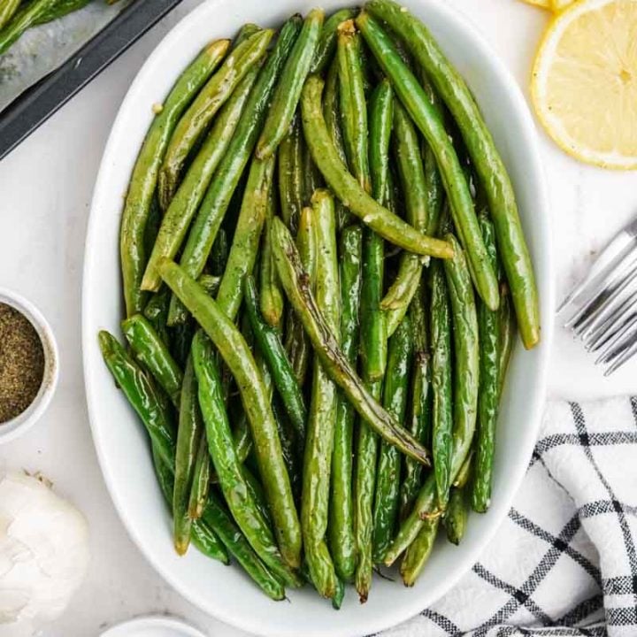 A shallow white dish of oven roasted green beans with additional seasonings around it.