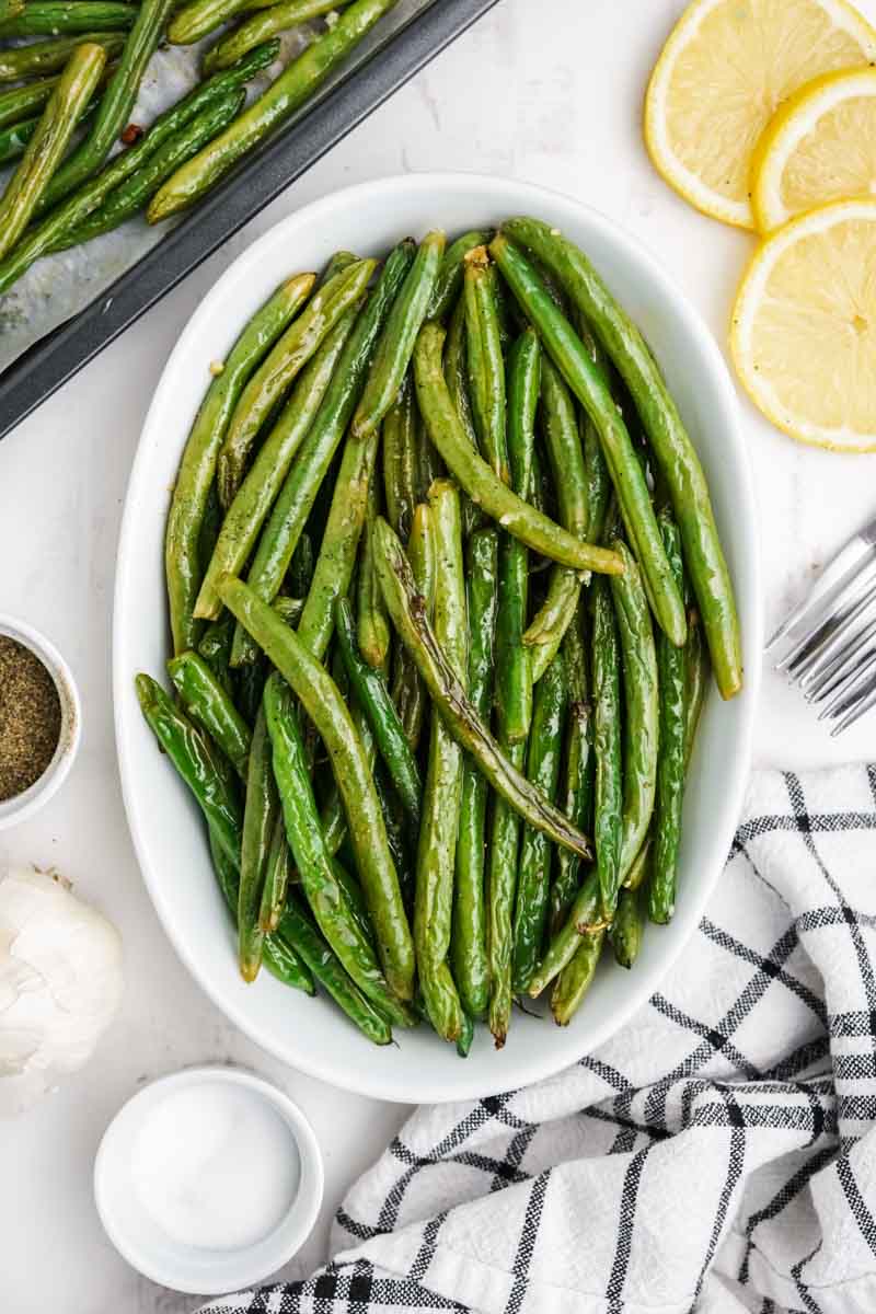 A shallow white dish of oven roasted green beans with additional seasonings around it.