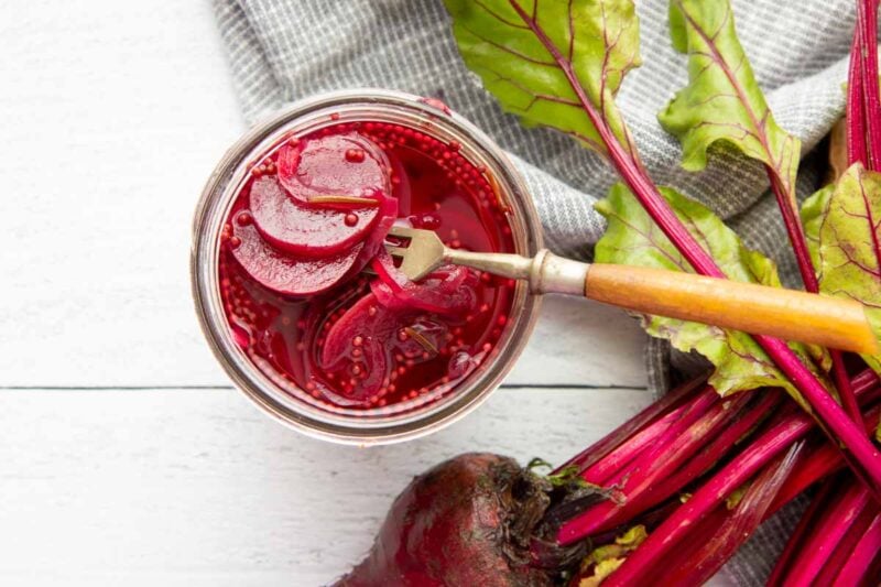 A three-pronged fork with a wooden handle gently lifts sliced beets from a flavorful brine in a canning jar.