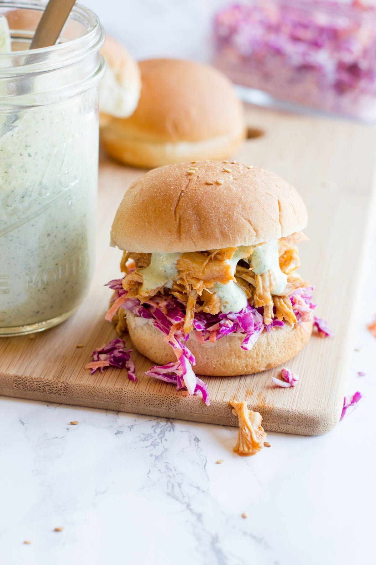 A slider topped with slaw, sriracha chicken, and ranch sauce sits atop a wooden cutting board on a marble counter.