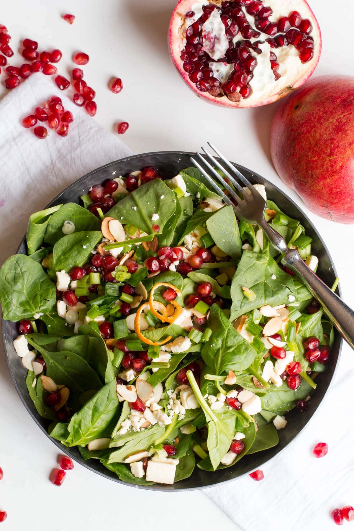 Overhead of spinach and pomegranate salad in a shallow bowl with pomegranate arils and halves alongside.