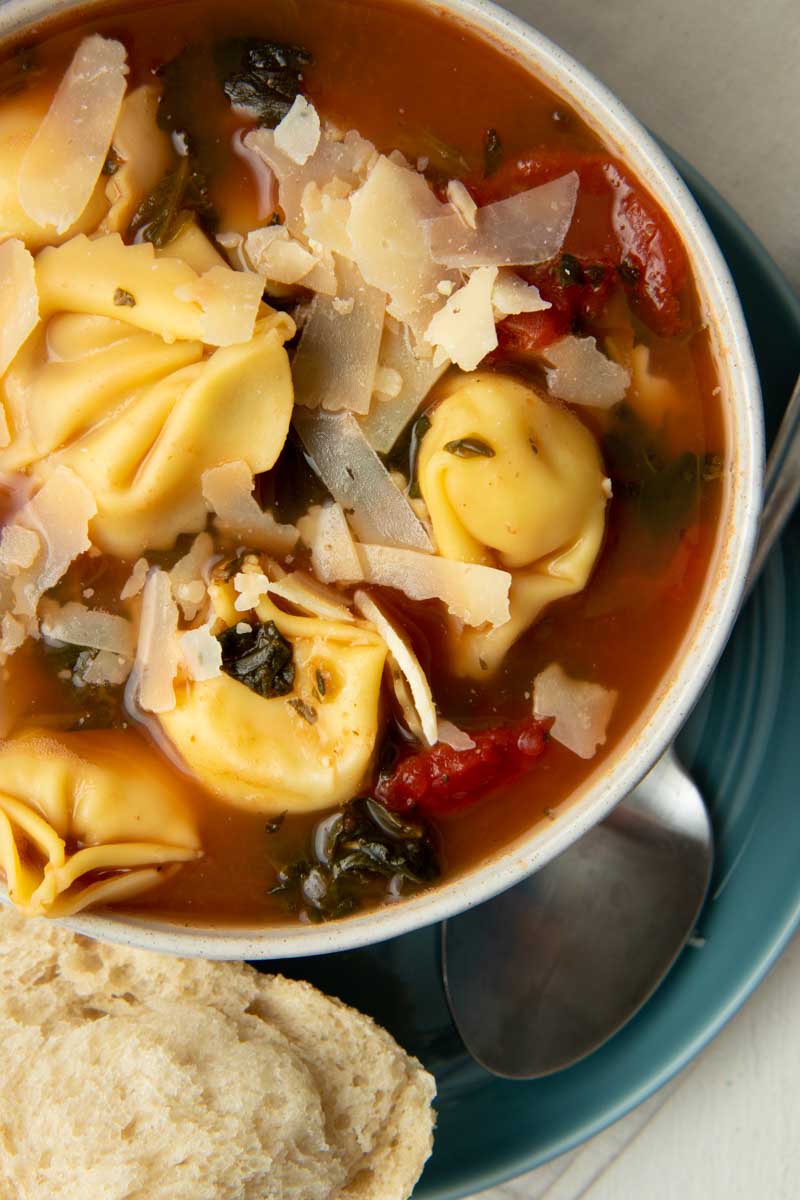 Close view of a bowl of spinach tortellini soup garnished with shaved parmesan, a spoon resting beside the bowl.